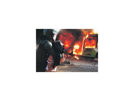 Una joven es detenida por un policía frente a un colectivo en llamas. La escena ocurrió en el Parque Bustamante, centro de Santiago.