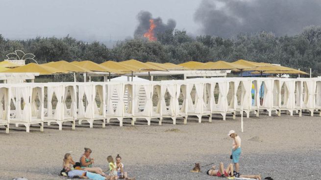 IRRUPCIÓN. La gente descansa en una playa mientras el humo y las llamas se elevan después de las explosiones en una base aérea militar rusa, en Novofedorivka, Crimea.
