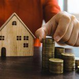 Una mano sostiene monedas y pequeñas casas de madera, símbolo del ahorro y del acceso al crédito hipotecario en un contexto de ajuste de tasas y mayores exigencias Una mano sostiene monedas y pequeñas casas de madera, símbolo del ahorro y del acceso al crédito hipotecario en un contexto de ajuste de tasas y mayores exigencias