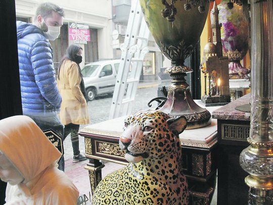 Vidriera. Desde afuera, dos personas miran el interior de una vidriera repleta de objetos antiguos y obras de arte.&nbsp;