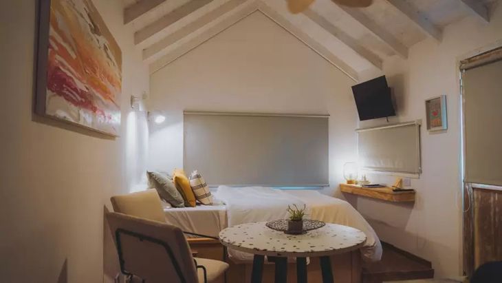 En un solo espacio se combinan cama matrimonial, mesa redonda y área de estar. Los techos de madera con tirantería a la vista, la iluminación puntual y los muebles flotantes completan una ambientación cálida En un solo espacio se combinan cama matrimonial, mesa redonda y área de estar. Los techos de madera con tirantería a la vista, la iluminación puntual y los muebles flotantes completan una ambientación cálida
