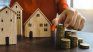 Una mano sostiene monedas y pequeñas casas de madera, símbolo del ahorro y del acceso al crédito hipotecario en un contexto de ajuste de tasas y mayores exigencias