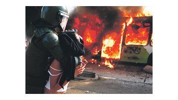 Una joven es detenida por un policía frente a un colectivo en llamas. La escena ocurrió en el Parque Bustamante, centro de Santiago.