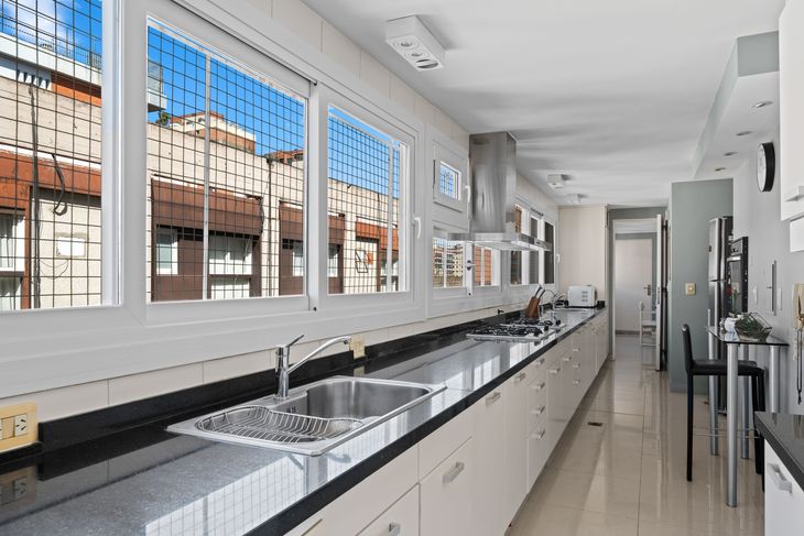 La cocina principal se extiende a lo largo del frente, con muebles blancos, mesada de granito y aberturas de aluminio con doble vidrio La cocina principal se extiende a lo largo del frente, con muebles blancos, mesada de granito y aberturas de aluminio con doble vidrio