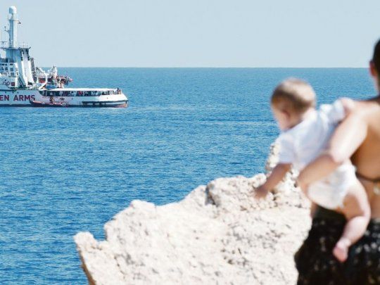 Contraste. Una turista con su bebé en brazos observa el barco con migrantes del Open Arms, a pocos metros frente a la costa de italiana de Lampedusa. Son 107 los refugiados que están a bordo desde hace 17 días.