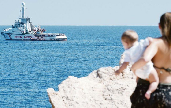 Contraste. Una turista con su bebé en brazos observa el barco con migrantes del Open Arms, a pocos metros frente a la costa de italiana de Lampedusa. Son 107 los refugiados que están a bordo desde hace 17 días.