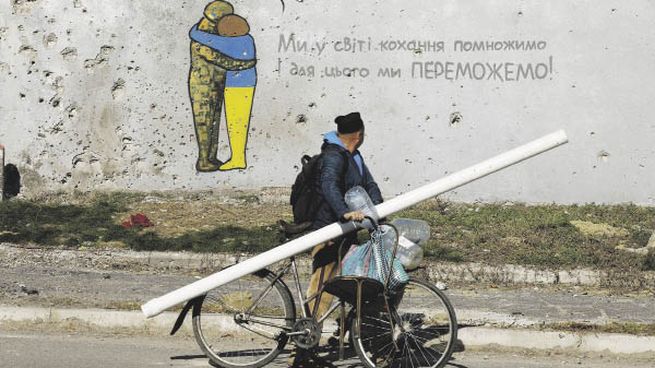 HUELLAS. Un hombre pasa en bicicleta frente a un grafiti en una pared de un edificio cubierto con rastros de balas y metralla, en la ciudad recientemente recuperada de Kupiansk, Ucrania.