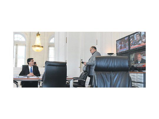 Jorge Capitanich duplicado: en su despacho con un colaborador mientras su imagen aparece en una de las pantallas de televisión.